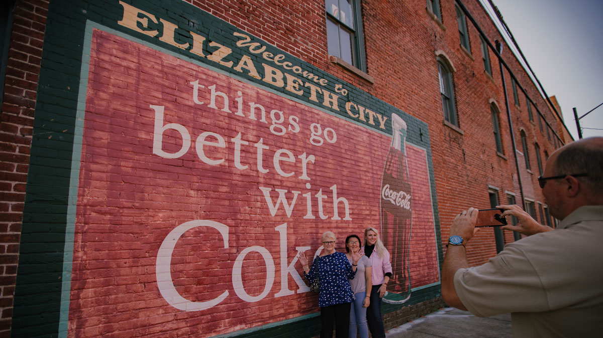 grandma, daughter and granddaughter smiling and posing for picture with coke mural in elizabeth city